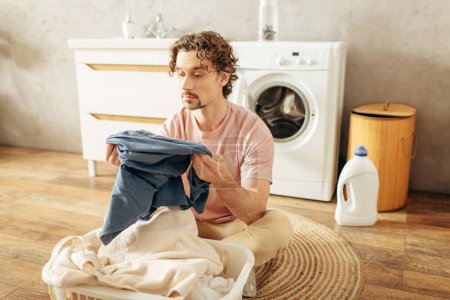 A handsome man in cozy homewear sitting next to a washing machine.