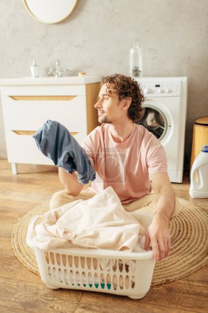 A handsome man in cozy homewear sitting beside a laundry basket.
