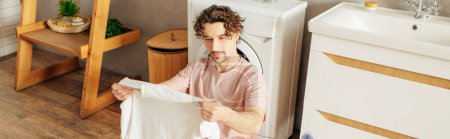 Foto de Un hombre guapo en ropa de casa acogedora está al lado de una lavadora en un baño. - Imagen libre de derechos