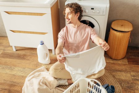 Un hombre guapo en ropa de casa acogedora sentado al lado de una lavadora.