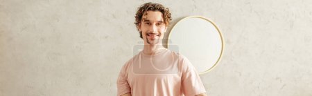 Un hombre guapo en ropa de casa acogedora se para frente a un espejo redondo.