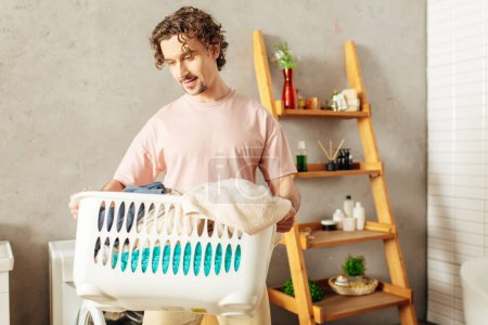 Ein gutaussehender Mann in gemütlicher Hauskleidung mit einem Wäschekorb im Badezimmer.
