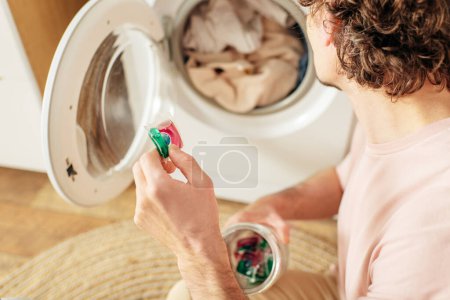 Foto de A man in cozy homewear holds a cup of detergent in front of a washing machine. - Imagen libre de derechos