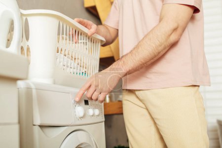 Handsome man in cozy homewear putting laundry basket on washing machine.