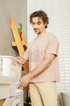 Un hombre guapo en ropa de casa acogedora de pie delante de una lavadora.
