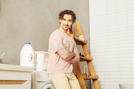 Foto de Un hombre guapo en ropa de casa acogedora de pie junto a una lavadora, listo para el día de la lavandería. - Imagen libre de derechos
