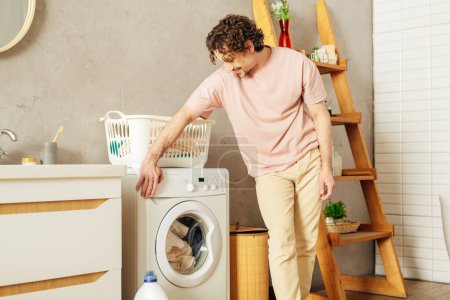Foto de A handsome man in cozy homewear stands next to a washing machine. - Imagen libre de derechos