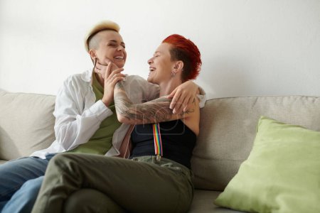 Lesbian couple with tattoos sitting closely on a sofa, showcasing their body art in a cozy indoor setting.