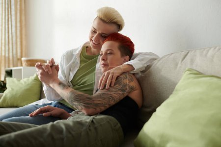 Two lgbt women with tattoos sit comfortably on a couch, sharing a moment of togetherness and self-expression at home.