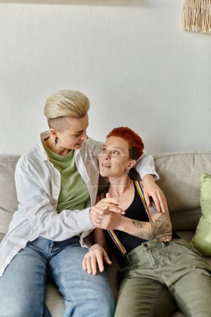 Foto de Dos mujeres, una pareja lesbiana con el pelo corto, están cómodamente sentadas juntas en un sofá en casa. - Imagen libre de derechos