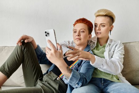 Two women with short hair sitting on a couch together, engrossed in a cell phone.