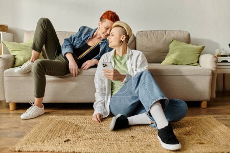 Téléchargez les photos : Un couple de lesbiennes aux cheveux courts assis sur le sol devant un canapé, profitant d'un moment de calme ensemble à la maison. - en image libre de droit