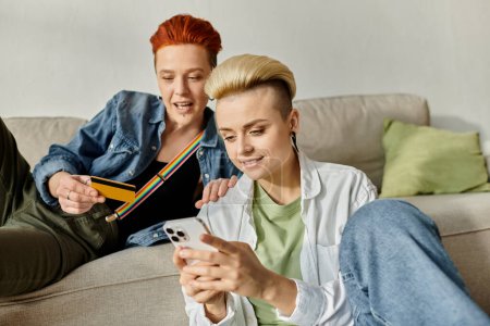 Téléchargez les photos : Deux femmes aux cheveux courts s'assoient sur un canapé, enveloppées dans un écran de téléphone portable, partageant un moment d'intimité et de connexion. - en image libre de droit