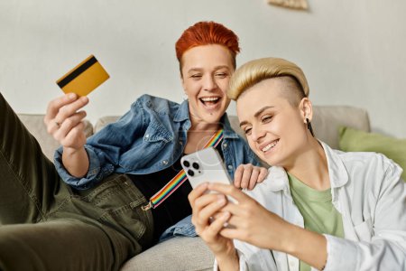 Foto de Dos mujeres, una pareja lesbiana con el pelo corto, se sientan en un sofá con una tarjeta de crédito, haciendo compras en línea juntas en casa. - Imagen libre de derechos