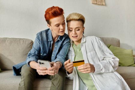 Foto de Una pareja lesbiana con el pelo corto se sienta en un sofá, examinando atentamente una tarjeta de crédito juntos. - Imagen libre de derechos