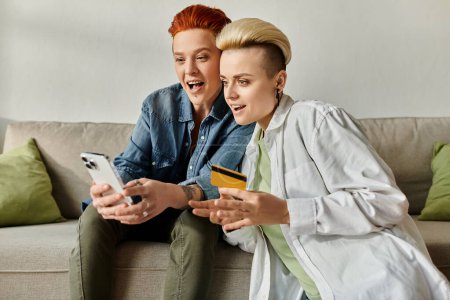 Two women with short hair sitting on a couch, contemplating a credit card in hand, in a cozy home setting.