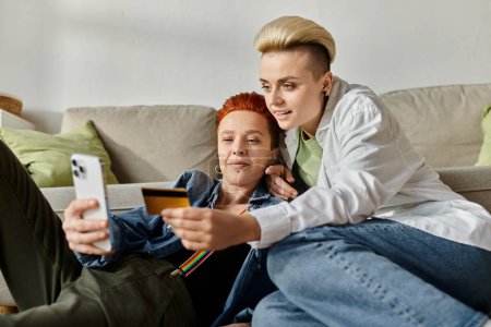Dos mujeres, una pareja lesbiana con el pelo corto, se sientan en el suelo en casa, discutiendo asuntos financieros mientras sostienen una tarjeta de crédito.