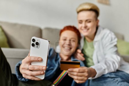 Foto de Una pareja lesbiana con el pelo corto sonríe mientras se toman una selfie juntos usando una tarjeta de crédito en casa. - Imagen libre de derechos