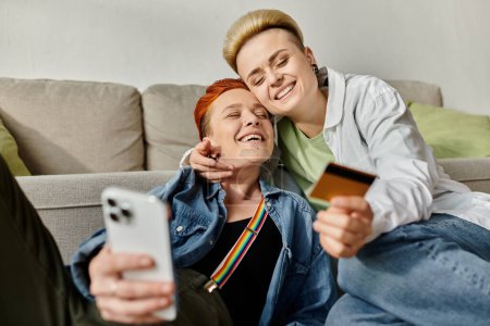 Zwei Frauen, ein lesbisches Paar, sitzen mit einer Kreditkarte in der Hand auf einer Couch und kaufen gemeinsam ein.