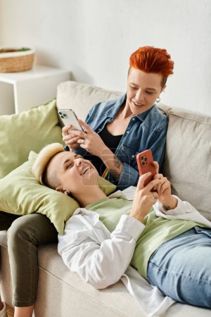Foto de Dos mujeres, una pareja lesbiana con el pelo corto, sentadas en un sofá absorto en sus teléfonos. - Imagen libre de derechos