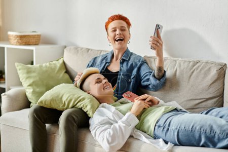 Dos mujeres, una pareja de lesbianas con el pelo corto, se sientan de cerca en un sofá, absortos en su teléfono celular.