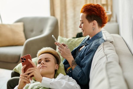 Foto de Dos mujeres con el pelo corto se sientan juntas en un sofá, absortas en sus teléfonos. - Imagen libre de derechos