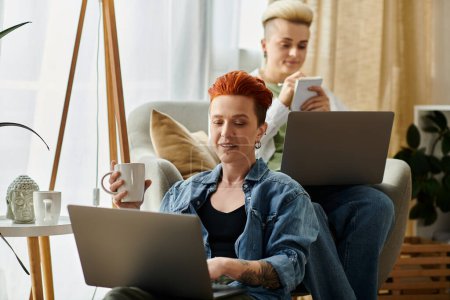 Dos mujeres con el pelo corto se sientan en un cómodo sofá, cada uno centrado en sus computadoras portátiles, absorto en su mundo en línea.