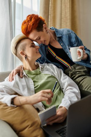Foto de Dos mujeres con cabello corto disfrutan del café y trabajan juntas en un portátil en un sofá, compartiendo un momento de conexión. - Imagen libre de derechos