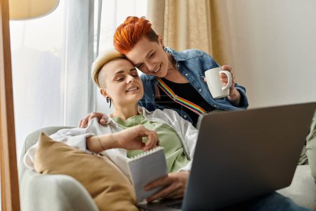 Zwei Frauen, ein lesbisches Paar mit kurzen Haaren, sitzen eng umschlungen in ihrem gemeinsamen Laptop auf einer Couch..
