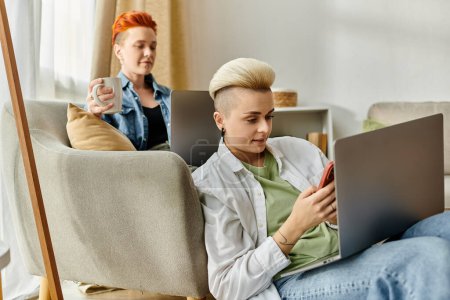 Dos individuos, una pareja lesbiana con el pelo corto, se sientan en un sofá usando computadoras portátiles en casa.