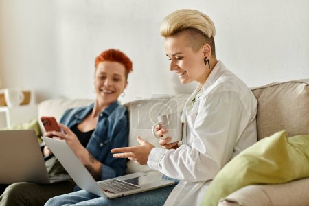 Photo for Two women, a lesbian couple with short hair, sit closely on a couch, each focused on their laptops, possibly working or sharing ideas. - Royalty Free Image