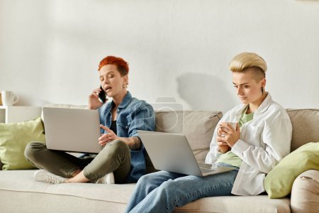 Foto de Dos mujeres con el pelo corto se sientan lado a lado en un sofá, cada uno absorbido en su propia pantalla del ordenador portátil. - Imagen libre de derechos