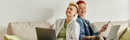 Lesbian couple with short hair engrossed in work on laptops while sitting closely on a couch at home.
