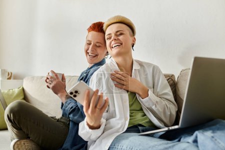 Téléchargez les photos : Deux femmes, un couple de lesbiennes aux cheveux courts, assises sur un canapé, concentrées sur leurs ordinateurs portables à la maison. - en image libre de droit