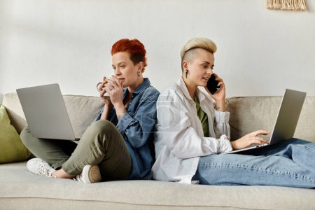 Foto de Una pareja lesbiana con el pelo corto se sienta en un sofá, cada uno absorto en su propio ordenador portátil. - Imagen libre de derechos