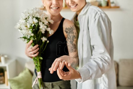 Two women hold bouquets of flowers in a cozy living room, showcasing engagement ring