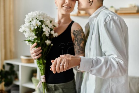 Foto de Lesbian couple holding flowers and showing wedding ring in a cozy living room. - Imagen libre de derechos