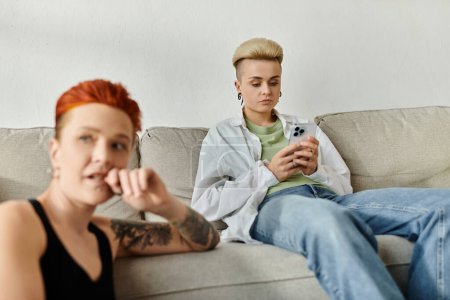 Foto de Dos personas, una pareja lesbiana con el pelo corto, se sientan en un sofá absorbido por el teléfono, desconectados el uno del otro. - Imagen libre de derechos