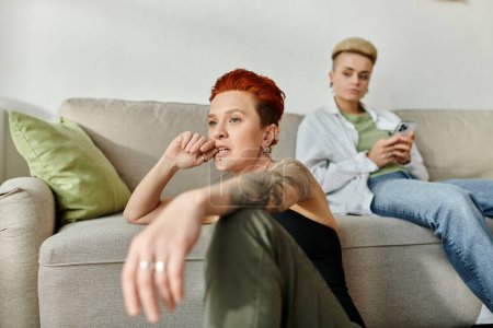 Two women with short hair sit next to each other, engrossed in separate activities while at home.