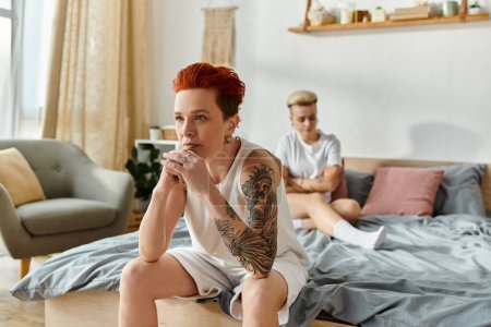 A lesbian couple with short hair sitting on a bed, showcasing their tattoos, having conflict