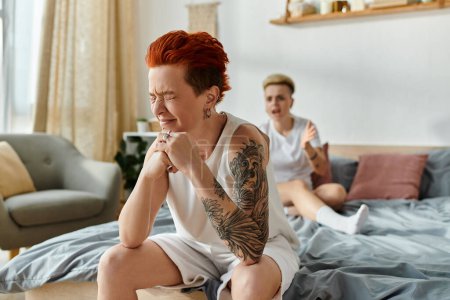 aufgebrachte Frau mit Tätowierung sitzt zusammen mit Partner und weint auf einem Bett in einem Schlafzimmer und präsentiert ihre einzigartige Körperkunst.