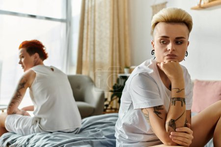 Two women with tattoos, a lesbian couple, sit together on a bed in a bedroom, showcasing relationship difficulties