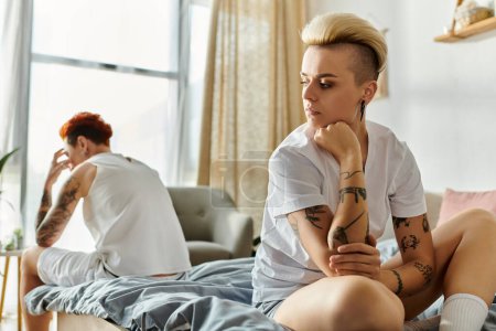 women, both with tattoos, sit closely on a bed in their bedroom, showcasing their unique sense of style and connection.