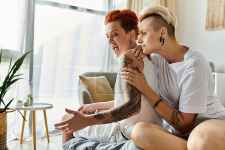 Two edgy women with tattoos sitting closely on a cozy couch in a stylish living room.