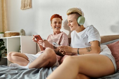 Two women with short hair sitting on a bed, engrossed in playing games together, showcasing a modern LGBT lifestyle.