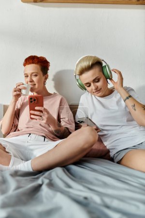 Foto de Dos mujeres, parte de un estilo de vida LGBTQ +, se sientan de cerca en una cama, uniéndose mientras escuchan música con auriculares. - Imagen libre de derechos