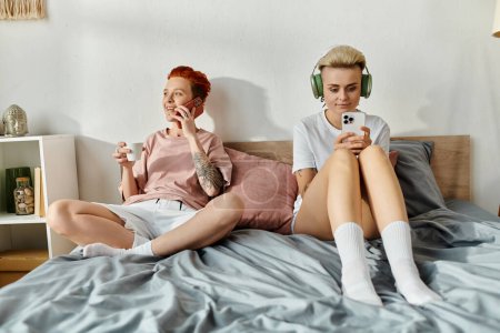 Photo for A lesbian couple with short hair enjoying music on headphones together in a cozy bedroom setting. - Royalty Free Image