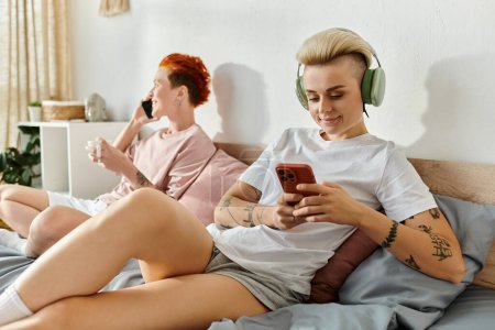 Téléchargez les photos : Deux personnes, un couple de lesbiennes, assis sur un lit et écoutant de la musique avec des écouteurs dans leur chambre. - en image libre de droit