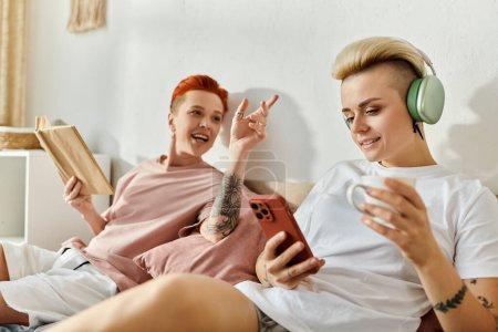 Foto de Una pareja lesbiana con el pelo corto se sienta en una cama, usando auriculares mientras disfrutan de la música juntos en su dormitorio. - Imagen libre de derechos