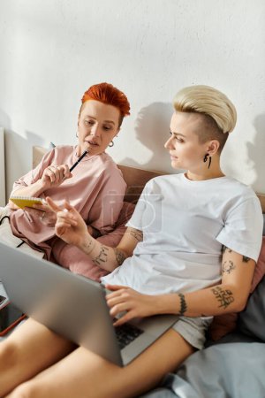Two women, a lesbian couple with short hair, sit on a bed and engage with a laptop, embodying a modern lgbt lifestyle.